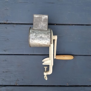 May include: A vintage white metal food grinder with a wooden handle and a silver metal container. The grinder has a crank handle and a clamp to secure it to a table.
