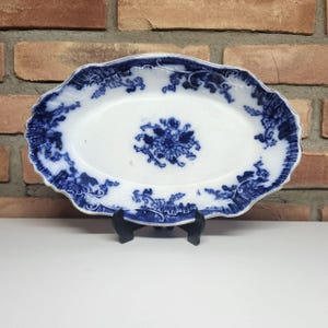 May include: Oval, antique serving platter with a white center and blue floral and scrollwork design. The rim has a scalloped edge. The platter is supported by a black stand. The background is a brick wall.