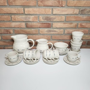 May include: A ceramic dinnerware set in cream with dark blue accents, including a large pitcher, a smaller creamer, four cups with matching saucers, a small bowl on a saucer, and a stack of three small bowls, all featuring a repeating floral pattern.