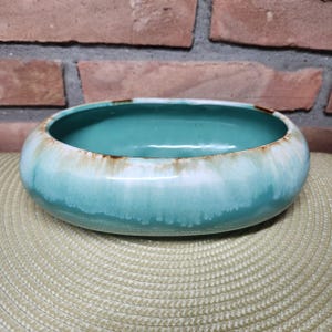 May include: Oval-shaped ceramic bowl with a turquoise interior and a light blue exterior. The rim has a white glaze with brown accents. The bowl is sitting on a beige woven mat, with a brick wall in the background.