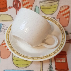 May include: A white teacup and saucer with a gold geometric pattern around the edge. The cup and saucer have a speckled white glaze.