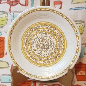 May include: A white ceramic dinner plate with a gold and brown floral design. The plate has a speckled texture and a raised rim.