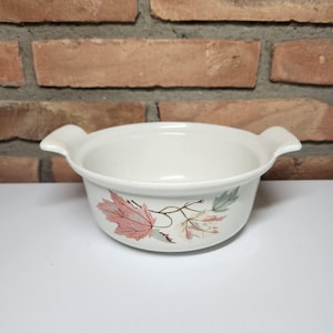May include: A white ceramic bowl with two handles, featuring a leaf design in pink, green, and brown. The bowl is round with a slightly flared rim, suitable for serving dishes.