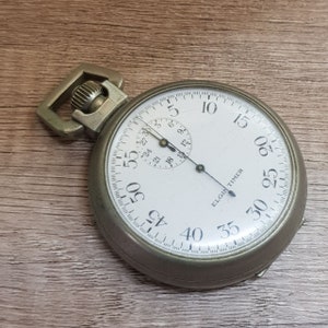 Op de afbeelding: Een zilveren zakhorloge met een witte wijzerplaat en zwarte markeringen. Het horloge heeft een zwarte wijzer en een kleinere wijzerplaat met zwarte markeringen. De wijzerplaat van het horloge geeft "ELGIN TIMER" aan.