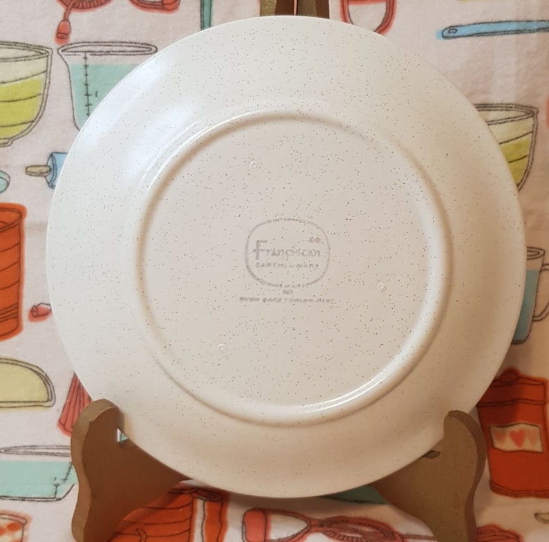 May include: White ceramic dinner plate with a speckled finish. The back of the plate has a circular logo that reads " Franciscan Co. Earthenware"