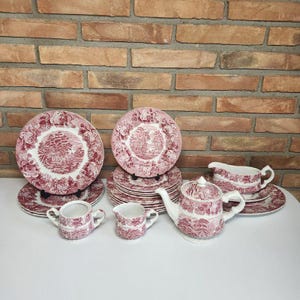 May include: A complete set of pink and white ceramic dinnerware with a scenic design. The set includes plates, saucers, a teapot, creamer, sugar bowl, and gravy boat. The dishes are arranged on a white surface against a brick wall.
