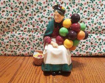 May include: A ceramic figurine of a woman wearing a green shawl and a red dress, holding a bunch of colorful balloons. She is sitting on a wooden surface and has a basket of flowers at her feet.