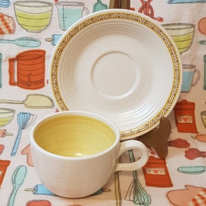May include: A white ceramic teacup with a yellow interior and a brown geometric pattern around the rim. The teacup is paired with a matching saucer with a brown geometric pattern around the rim.
