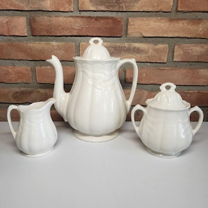 May include: A white porcelain teapot, creamer, and sugar bowl set. The teapot has a rounded body with a floral design. The creamer and sugar bowl are also decorated with a floral design.