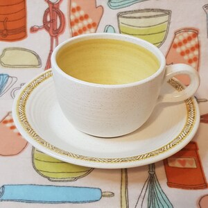 May include: A white ceramic teacup and saucer with a gold trim. The cup has a speckled finish and is filled with a yellow liquid. The saucer has a decorative pattern around the edge.