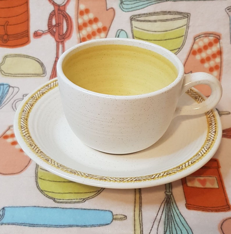 May include: A white ceramic teacup and saucer with a gold trim. The cup has a speckled finish and is filled with a yellow liquid. The saucer has a decorative pattern around the edge.