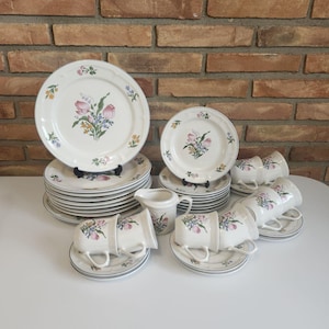 May include: A set of white ceramic dinnerware with a floral design featuring pink, yellow, and green flowers. The set includes dinner plates, salad plates, bowls, cups, and a creamer.