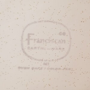 May include: Close-up of a white ceramic plate with a brown speckled glaze. The plate has a circular logo that reads "Franciscan Co. Earthenware" in a gray font.