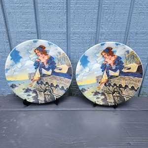 May include: Two decorative ceramic plates with a blue and white design featuring a woman in a blue dress standing on a stone ledge overlooking a body of water. The plates are on black stands.