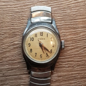 May include: A vintage silver Timex wristwatch with a cream-colored face and black hands. The watch has a metal band and a round case.