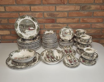 May include: A large ceramic dinnerware set featuring a winter landscape scene with a red barn and trees. The collection includes multiple stacks of dinner plates, salad plates, bowls, and teacups with matching saucers, all decorated with a green border.