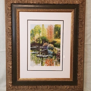 May include: Framed print of a colorful landscape painting with a reflection of trees in a still body of water. The print is signed "Claude Monet" and dated "1903".