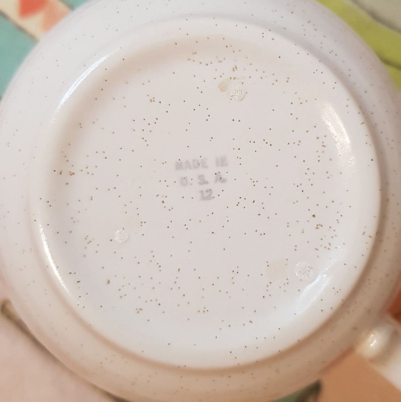 May include: White ceramic mug with a speckled glaze. The bottom of the mug has the text "Made in U.S.A. 12" printed in black.