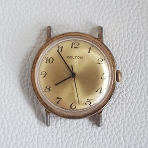 May include: A gold-toned Kelton wristwatch with black hands and black Roman numeral hour markers on a gold-colored face. The watch has a silver-toned metal band.