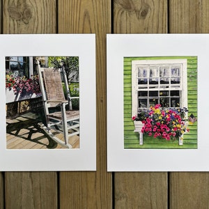 May include: Two framed watercolor paintings. One depicts a porch with a rocking chair and flower boxes. The other shows a window with a flower box and an American flag. The paintings are on a wooden background.