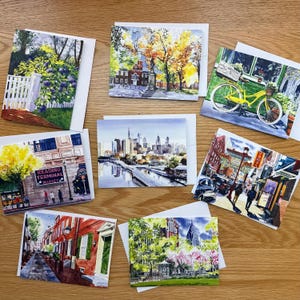 May include: A collection of watercolor art greeting cards featuring cityscapes and landscapes. The cards depict scenes such as a yellow bicycle, a city skyline, and a street scene with the text "Reading Terminal Market". Each card is paired with a white envelope.
