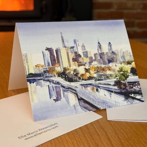 May include: Watercolor greeting card featuring a cityscape with tall buildings, a bridge, and water reflections. The artwork showcases a variety of colors, including blues, browns, and yellows. The card is folded and accompanied by a white envelope. Text on the card reads "Ellie Moniz Watercolors".