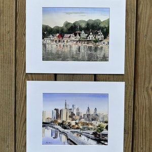 May include: Two framed watercolor paintings. The top painting depicts a row of boathouses along a river, with a backdrop of green trees and a blue sky. The bottom painting shows a city skyline with tall buildings and a bridge.