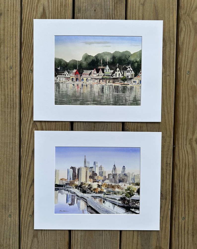 May include: Two framed watercolor paintings. The top painting depicts a row of boathouses along a river, with a backdrop of green trees and a blue sky. The bottom painting shows a city skyline with tall buildings and a bridge.