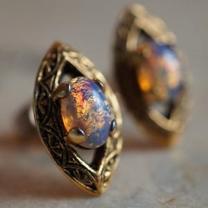 May include: A pair of vintage-style stud earrings. Each earring features an oval, iridescent gemstone set within a gold-toned, filigree-detailed frame. The gemstones display a mix of orange, blue, and yellow hues, creating a shimmering effect. The earrings are displayed on a light-colored surface.