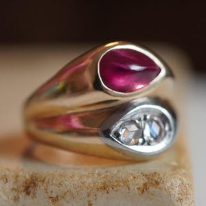 May include: A gold ring with two teardrop-shaped settings. The top setting holds a vibrant red gemstone, while the bottom setting features two smaller, clear gemstones. The ring is set on a textured, light brown surface.
