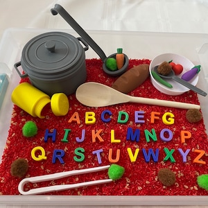 May include: A clear plastic bin filled with red rice, toy cooking utensils, and colorful alphabet letters. Includes a gray pot, wooden spoon, and toy vegetables. Educational and sensory play set.