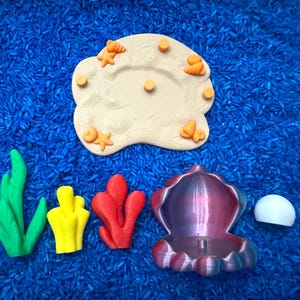 colorful ocean-themed toys, including a beige island with starfish and orange accents, green and yellow seaweed, red coral, a purple and blue shell, and a white pearl that piece together.
