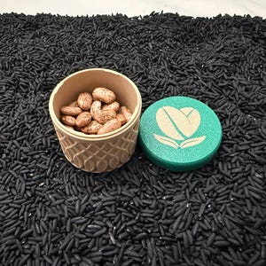 May include: A beige, patterned container filled with brown beans, next to a teal lid with a white coffee bean heart logo. The objects rest on a bed of black rice.