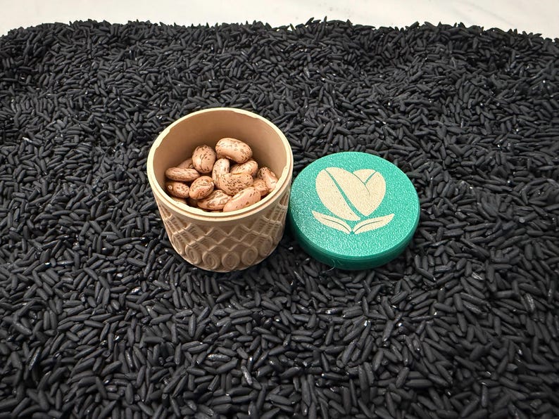 May include: A beige, patterned container filled with brown beans, next to a teal lid with a white coffee bean heart logo. The objects rest on a bed of black rice.