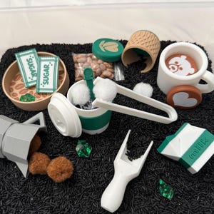 May include: A collection of toy coffee shop items, including a white mug with a bear design, a miniature coffee maker, and sugar packets. The items are arranged on a bed of black rice, with green and brown accents.