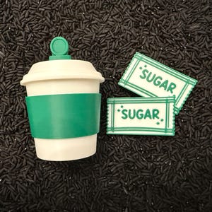 May include: A white and green coffee cup with a green sleeve and lid, next to two sugar packets. The sugar packets are white with green borders and the word "SUGAR" printed on them.
