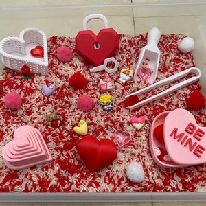 May include: A white plastic storage container filled with red and white rice, pink and white pom-poms, and various Valentine's Day themed toys, including a heart-shaped lock, a heart-shaped box that says "Be Mine", and a set of nesting hearts.
