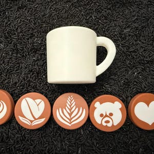 May include: A white ceramic mug with a curved handle sits above five round, brown coasters. Each coaster features a different white latte art design: a heart, coffee bean, fern, bear, and heart.