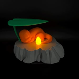 May include: An orange figurine of a creature resting under a green leaf, atop a gray, rocky base. A yellow light emanates from the center, creating a warm glow. The design is a stylized representation of a popular character.
