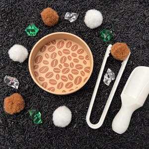 May include: A sensory play set with a round wooden tray featuring a coffee bean design. The set includes brown pom-poms, white tongs, a white scoop, clear and green gems, and white pom-poms, all arranged on a bed of black rice.