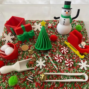May include: A Christmas sensory bin with red and green rice, a snowman, a Christmas tree, a red sleigh, a white scoop, a red gift box with a green bow, candy canes, and white snowflakes.