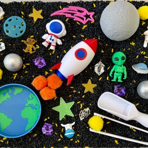 May include: A collection of space-themed toys and sensory items, including a white and red rocket, astronauts, a green alien, a moon, stars, and a planet Earth. The items are arranged on a black surface.