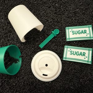 May include: A disassembled coffee cup set with a white cup, green sleeve, and lid. A green stirrer and two sugar packets with "SUGAR" text are also present. The items are on a black surface.