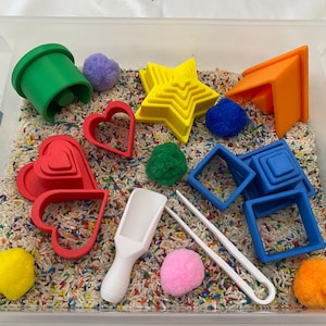 May include: A clear plastic bin filled with sensory materials. Includes colorful shapes like hearts, stars, and squares, along with pom-poms, a scoop, and tongs. The base is filled with rice and colorful sprinkles. A fun, educational toy.