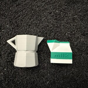 May include: A gray, geometric coffee maker and a white and green milk carton are displayed on a bed of black rice. The milk carton has the word "milk" printed on it. The objects are likely decorative.