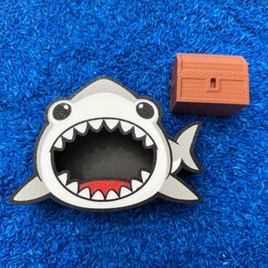 A shark-shaped container with a wide open mouth, black interior, and red accents. A small, brown treasure chest is next to the shark. The objects are on blue rice.
