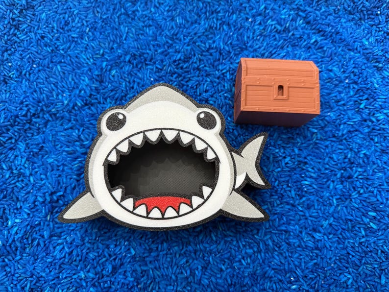 A shark-shaped container with a wide open mouth, black interior, and red accents. A small, brown treasure chest is next to the shark. The objects are on blue rice.