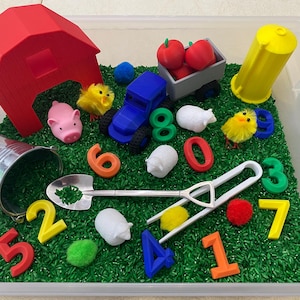 May include: A red barn, a blue toy truck, a bucket, a shovel, and a variety of farm animals are placed in a clear plastic bin filled with green rice.  The numbers 1 through 8 are scattered throughout the bin.