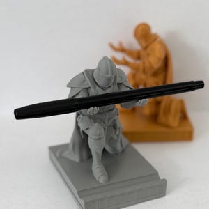 May include: A gray knight figurine holding a black pen, kneeling on a gray base. The knight is wearing armor and a helmet. Behind the knight is a smaller, brown figurine of a person kneeling.