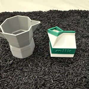 May include: A gray, geometric coffee maker and a white milk carton with green accents and the word "milk" printed on it. The objects are set against a backdrop of black rice.